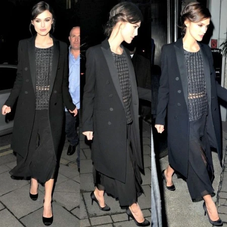 Keira Knightley wearing a black double-breasted coat, a patterned top, and flowing black pants. She is seen on a city street at night.