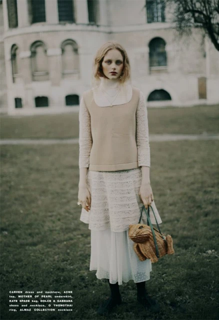 A woman wearing a long white dress, a tan vest, and holding a frog-shaped purse stands in a grassy field in front of a building.