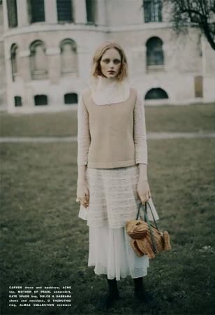 A woman wearing a long white dress, a tan vest, and holding a frog-shaped purse stands in a grassy field in front of a building.