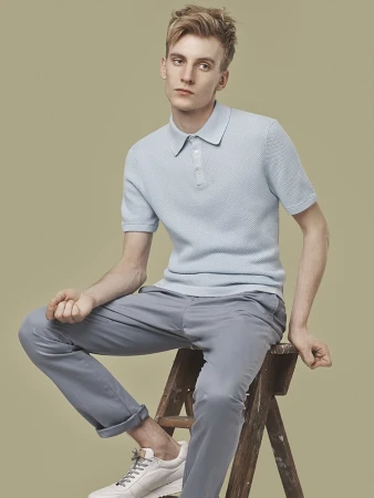 Fashion portrait of a blond male model wearing a light blue polo shirt and grey pants sitting on a wooden stool in a studio.