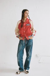 Model with braided hair wearing a red and cream varsity jacket and blue jeans against a white brick wall