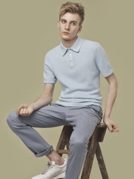 Young man in light blue polo shirt and grey trousers seated on a rustic wooden stool against a muted background