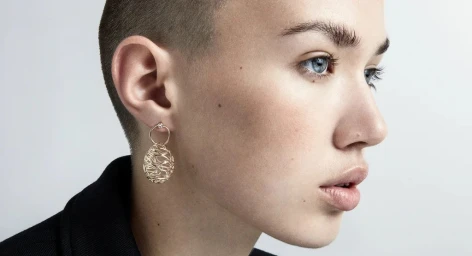 Close-up profile of a woman with a buzzcut wearing a gold wirework drop earring against a seamless light grey background