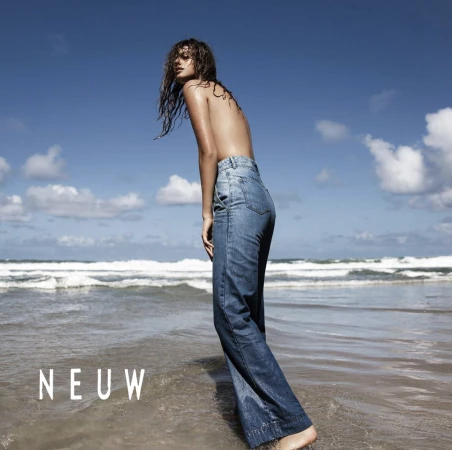 Model with wet hair wearing blue denim jeans standing in shallow ocean water at a sandy beach under a blue sky.