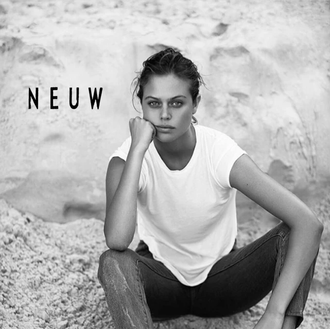 Black and white portrait of a young woman in a white t-shirt and jeans, sitting on a sandy surface with the word 'NEUW' in the background.