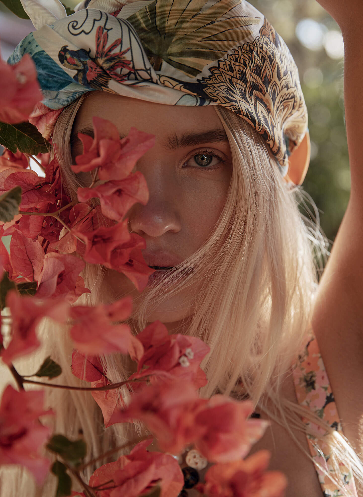 Close-up portrait of a woman with blonde hair wearing a floral scarf, posing with pink bougainvillea flowers in an outdoor setting.