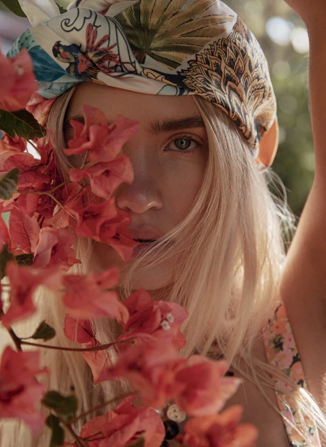 Close-up portrait of a woman with blonde hair wearing a floral scarf, posing with pink bougainvillea flowers in an outdoor setting.