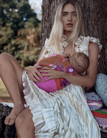 Woman sitting under tree breastfeeding baby wrapped in blanket, wearing white dress and floral embellishments, outdoor setting with green backdrop.