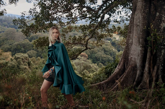 Blonde woman wearing a glossy green satin cape standing beside a large tree trunk in a lush forest landscape.