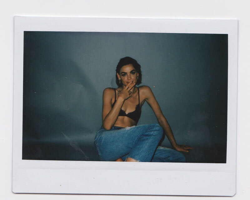 Portrait of a woman with short dark hair wearing a black bra and ripped jeans, seated against a grey backdrop in an indoor setting.