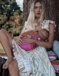 Blonde woman wearing a pleated white dress holding a baby in a patterned blanket outdoors against a tree trunk