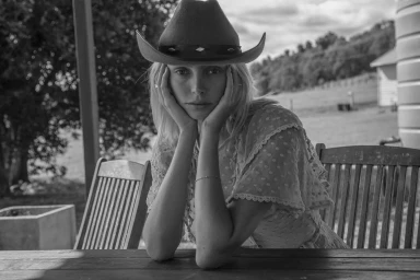 Monochrome portrait of a woman wearing a cowboy hat with hands cupping her face, seated outdoors.
