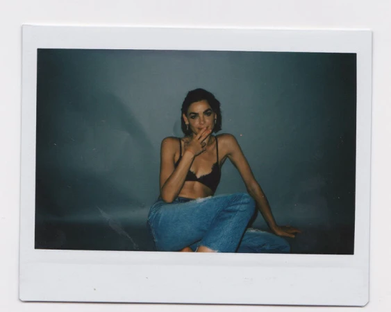 Polaroid photograph of a woman with dark hair wearing a black bra top and blue denim jeans against a grey background.