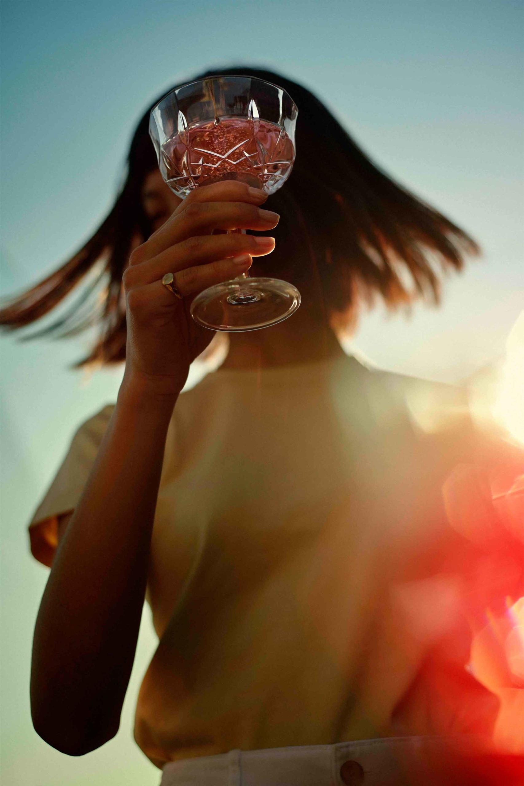 Woman holding a crystal wine glass with pink liquid against a clear blue sky with sun flare and wearing a yellow top.