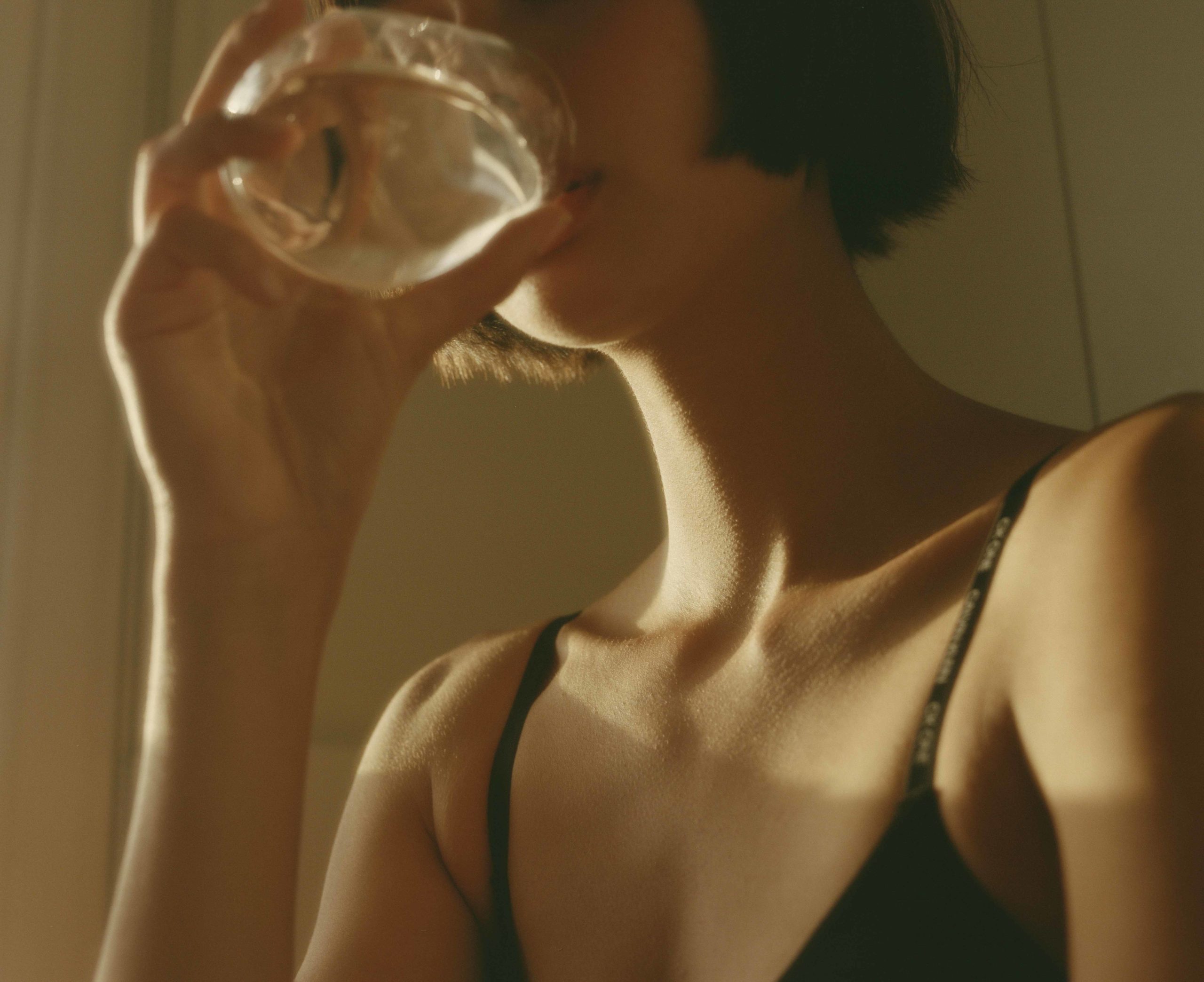 Close-up of a woman drinking water from a clear glass indoors with soft, diffused sunlight creating warm highlights on her skin.