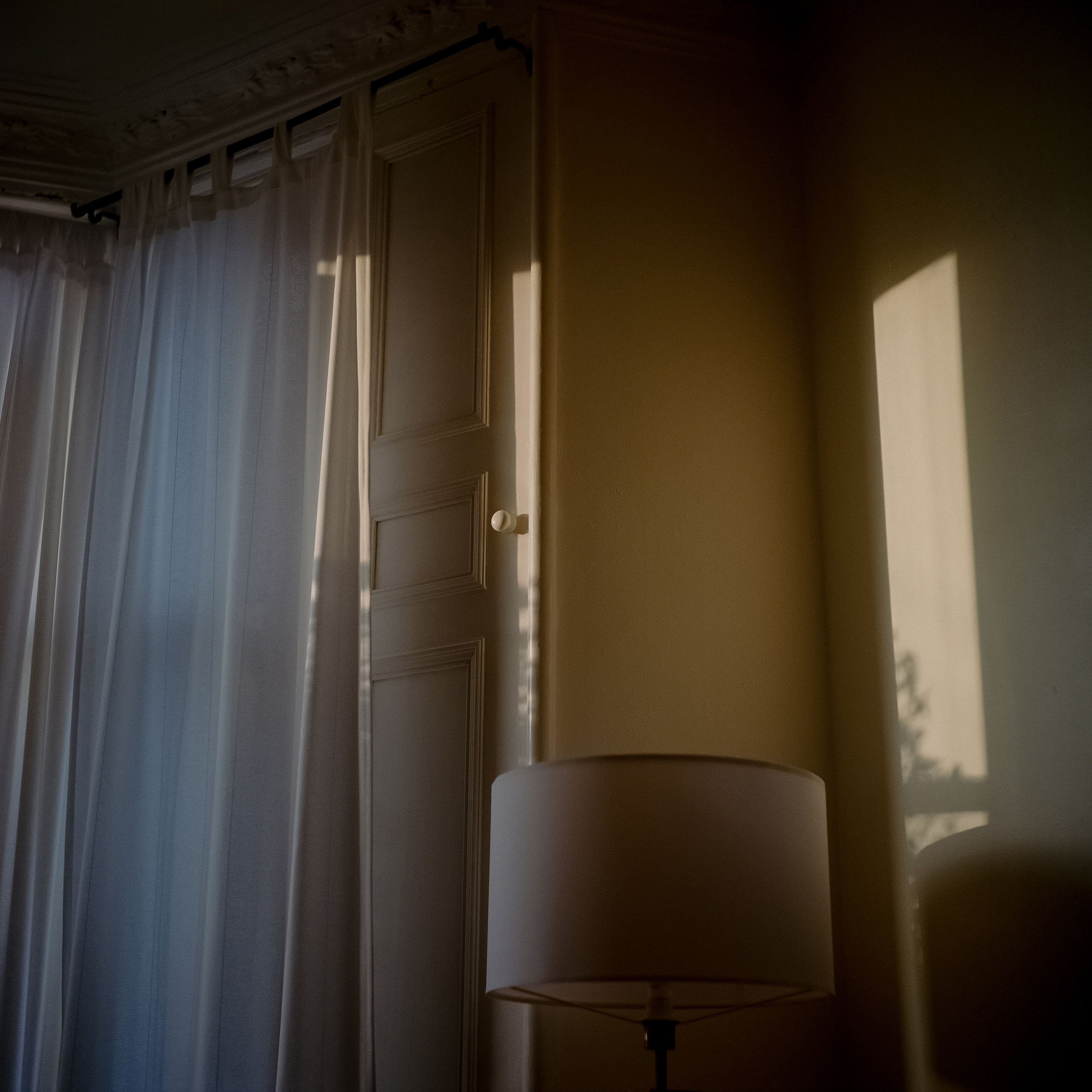 Interior view showing sheer white curtains, wooden shutters, and a lampshade lit by natural afternoon sunlight.