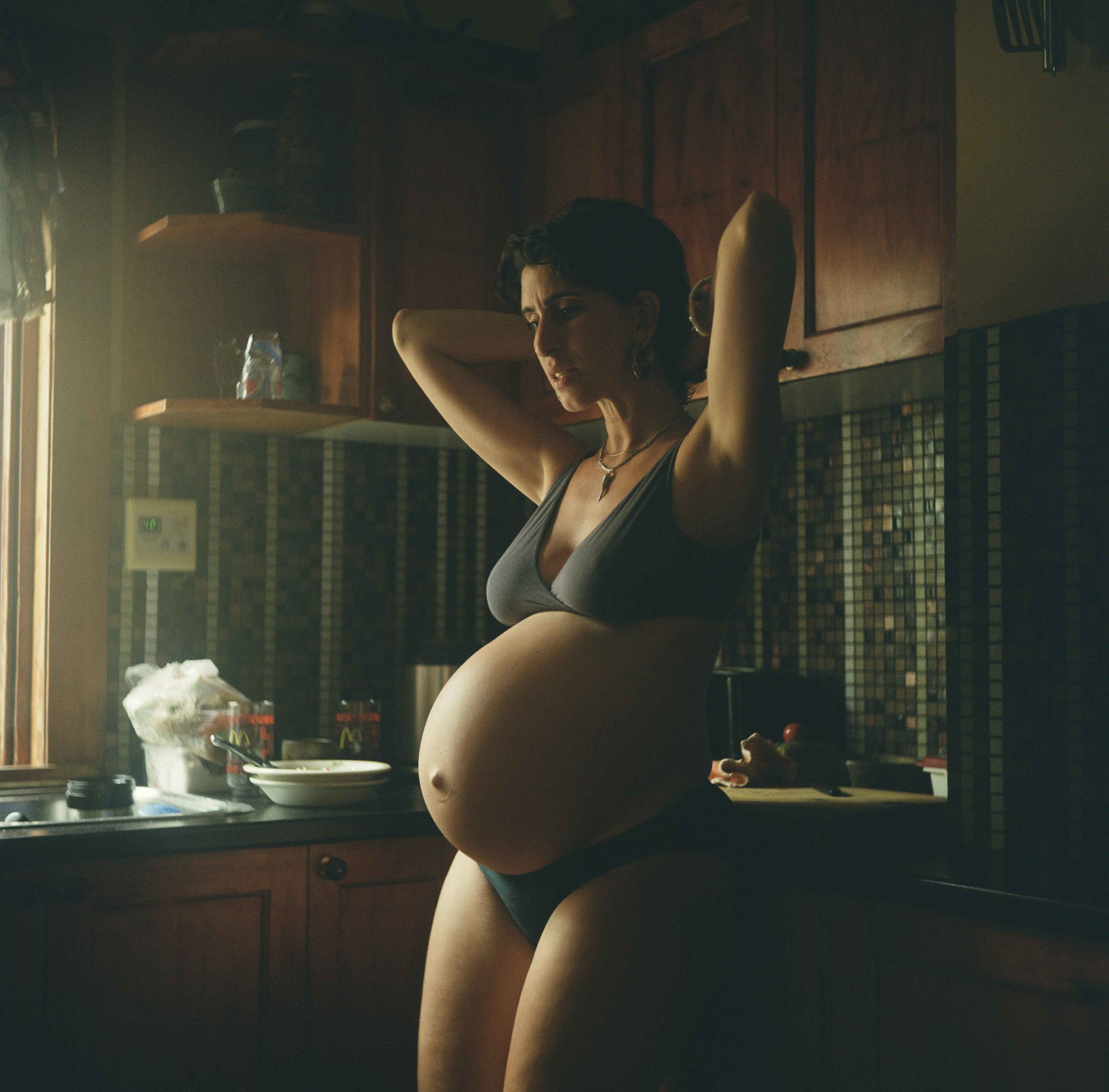 Pregnant woman in underwear stands in a kitchen with tiled backsplash and wood cabinets in a dimly lit setting with window light.