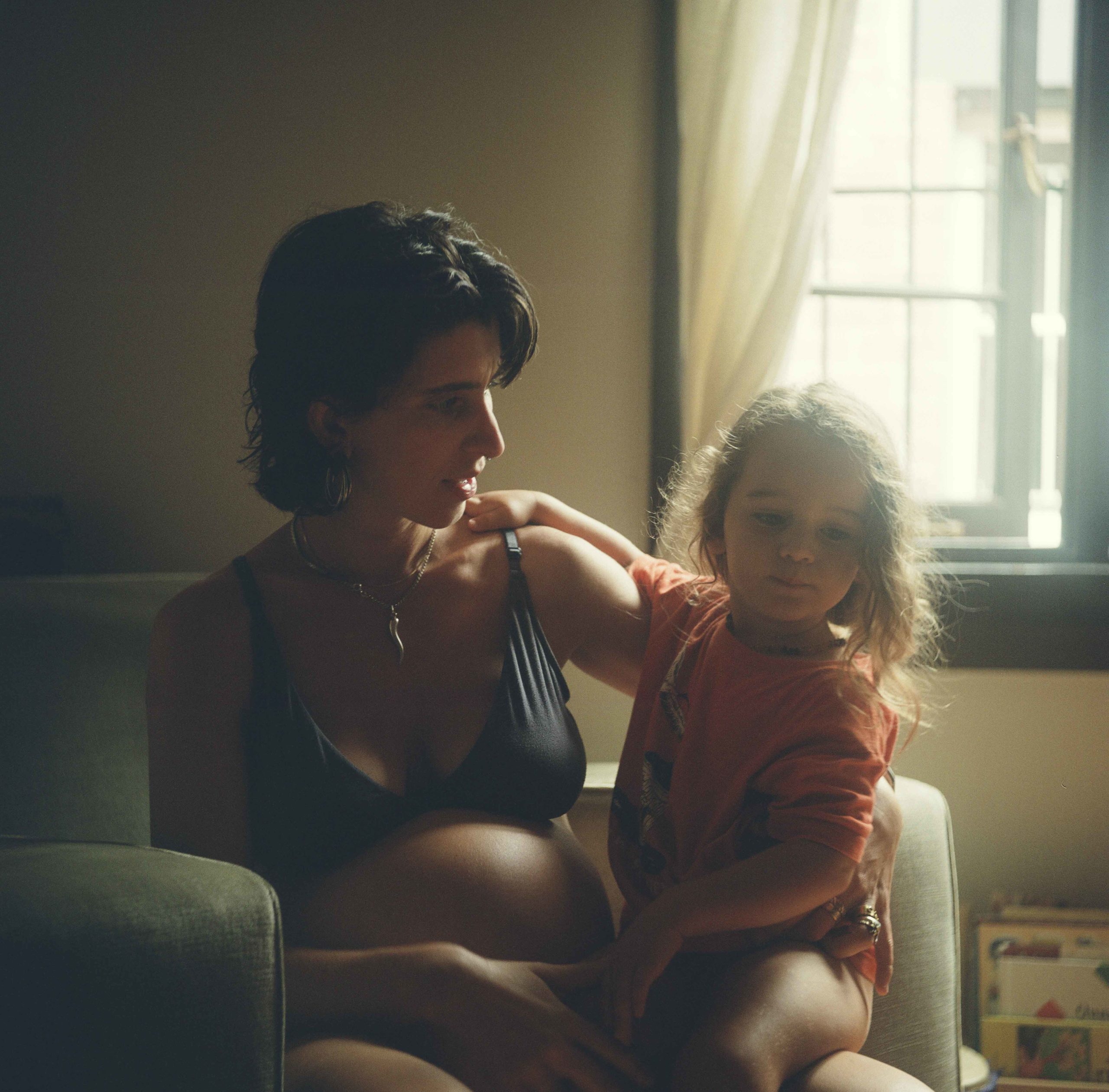 Pregnant woman sits in a chair in an indoor environment with a child on her lap, looking at her with a neutral expression.