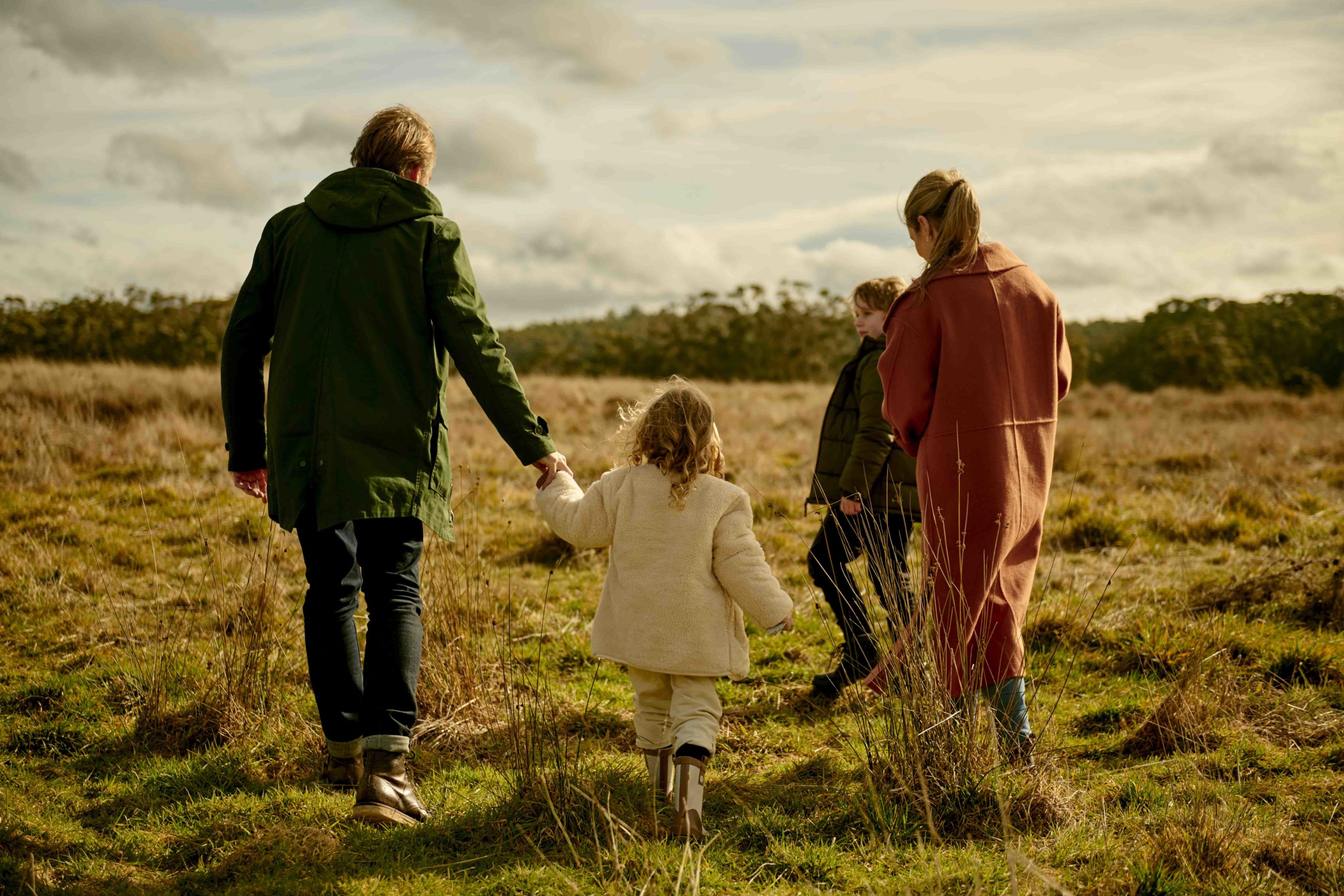 Family of four walks together through a grassy field. A young girl holds hands with her father in the foreground, and her brother and mother follow.