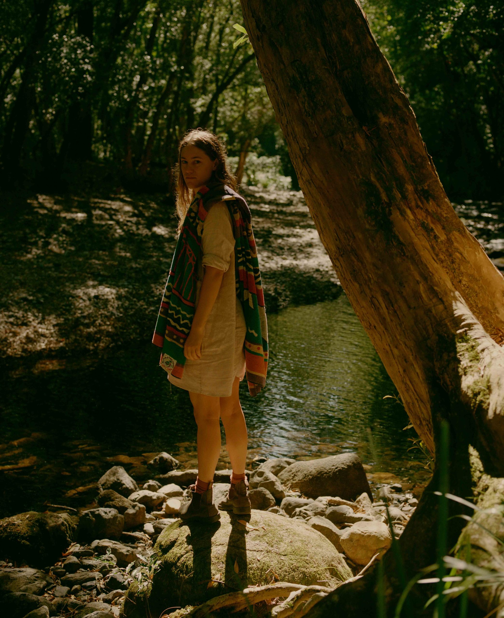 Woman stands on mossy rocks near a stream in a sun-dappled forest. She wears a light dress and a colorful shawl, looking over her shoulder.