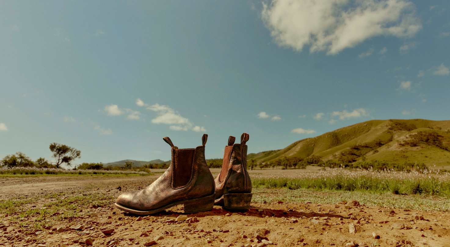 Two weathered leather boots rest on dry earth beneath a sunny, partly cloudy sky and rolling green hills in an outdoor rural setting.