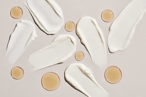 Overhead shot of white cream smears and golden gel droplets on a light beige surface, showcasing beauty or skincare product textures.