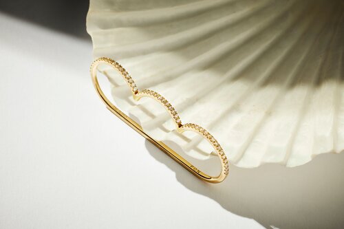 Close-up of a gold three-finger ring with gemstone detailing, set against a textured, off-white surface. Jewellery product shot.