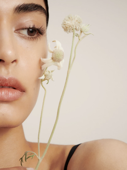 Close-up portrait of a woman with light skin, holding white flowers, set against a neutral background, focusing on beauty and natural elements.