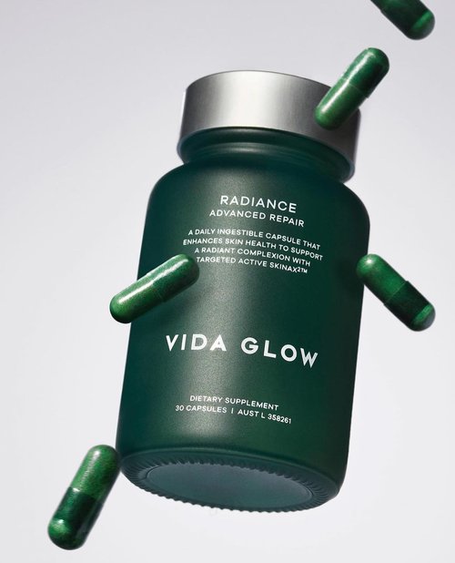 Bottle of Vida Glow dietary supplement capsules with surrounding green capsules against a grey background in a studio shot.