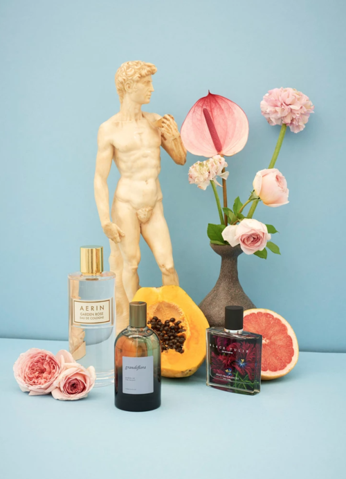 Still life composition featuring perfume bottles, a David statue replica, tropical flowers, and a grapefruit half on a light blue surface.