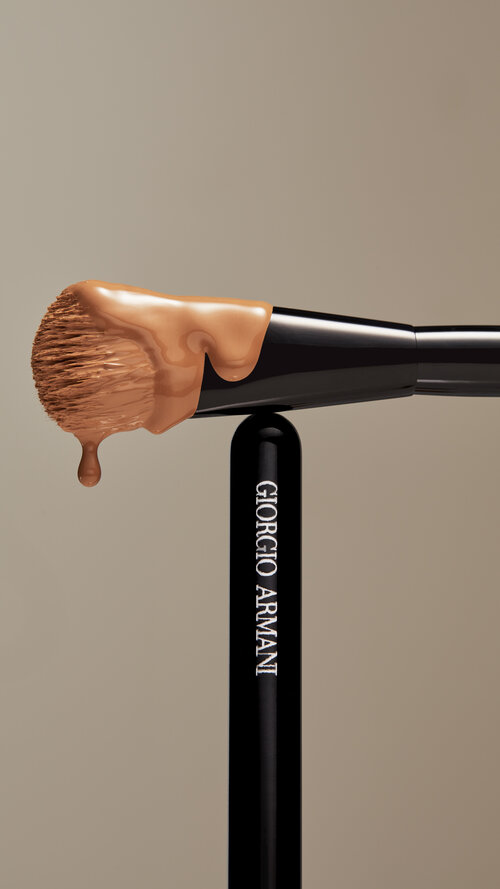 Cosmetic ad featuring makeup brush with foundation on the bristles and Giorgio Armani logo along the handle against a tan backdrop.