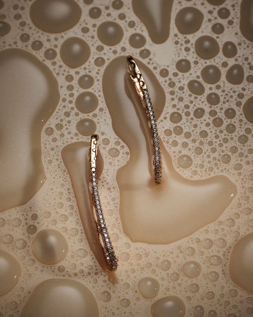 Elevated view of two gold and diamond hoop earrings placed on a light-beige surface with liquid-like bubbles creating a textured background.