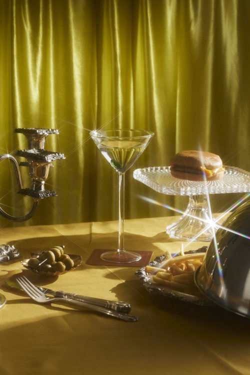 Elegant food still life featuring a martini, burger, and olives on a golden-yellow table with a matching draped curtain backdrop.