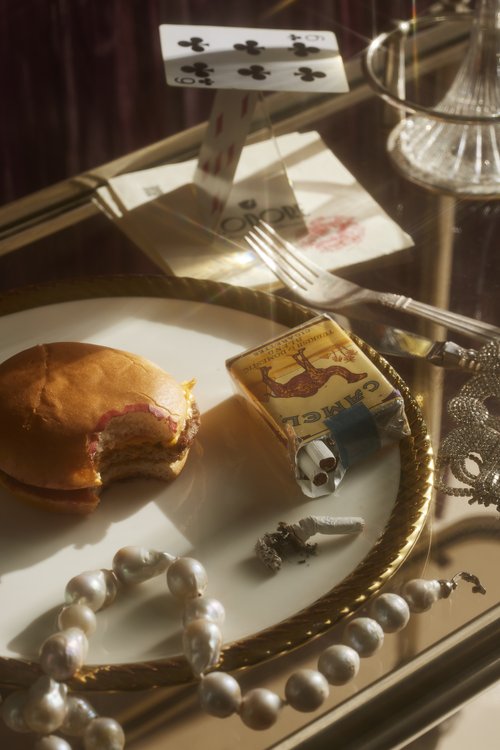 Still life photograph features a burger, playing cards, pearls, and cigarettes arranged on a mirrored gold tray, casting soft light.
