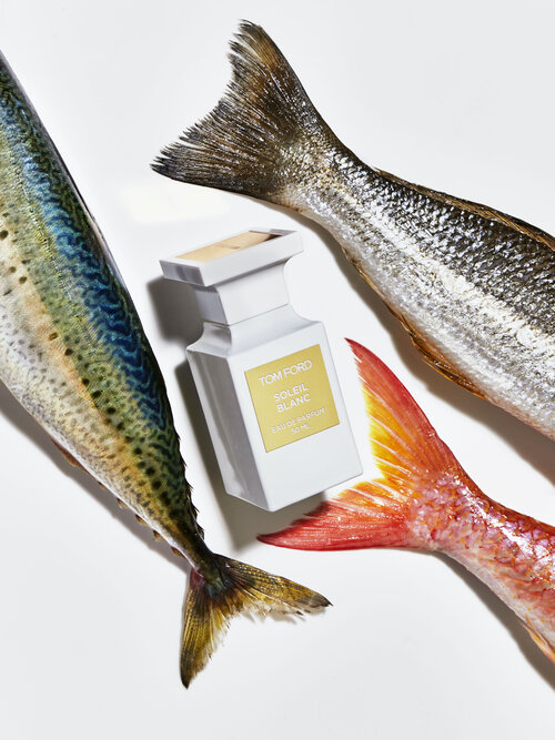Tom Ford Soleil Blanc perfume bottle surrounded by three fish tails in a studio setting with bright, even lighting.