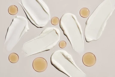 Overhead view of white cream smears and amber oil droplets scattered across a neutral beige background