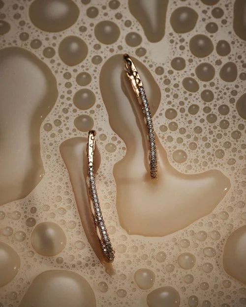 Two curved diamond and gold earrings resting among soapy water bubbles and foam.
