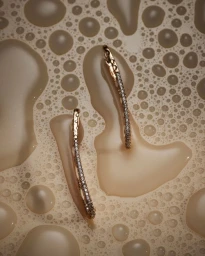 Two curved diamond and gold earrings resting among soapy water bubbles and foam.