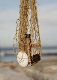 Beauty products and cosmetics suspended inside a woven string net bag against an out of focus coastal background.