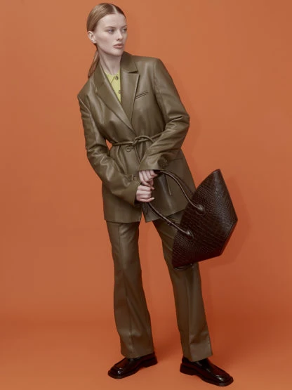 A fair-skinned adult woman poses in a leather suit, holding a dark brown woven tote bag against an orange seamless background.