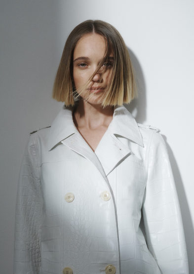 Close-up portrait of a young adult woman with blonde bob hairstyle wearing a light-colored double-breasted textured trench coat in a photo studio.
