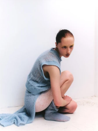 Woman in blue knitwear poses in a crouched position against a white wall in a fashion shot with yellow eye makeup and a slight frown.