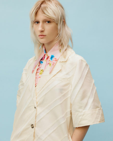 Blonde-haired woman in a cream-colored jacket over a floral shirt gazes directly at the camera against a blue background in a studio shot.