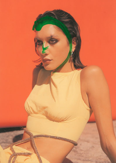 Young woman with green painted streaks across her face wears a yellow crop top against an orange background in a fashion-style photograph.