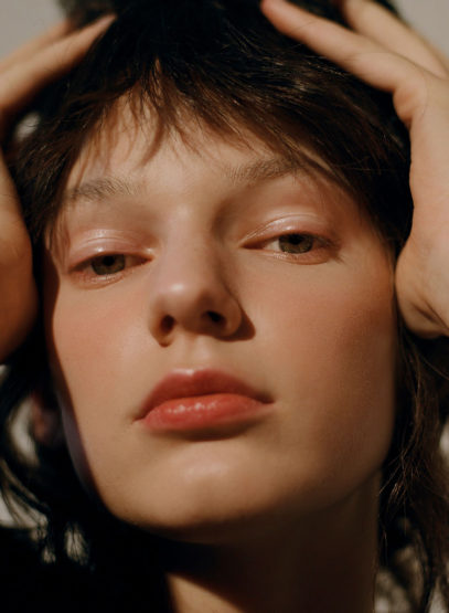 Close-up beauty shot of a young woman with fair skin, her hands placed on her head, soft lighting highlighting her features.