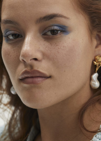 Close-up portrait of a woman with blue eyeshadow and decorative gold and pearl earrings, showcasing a beauty or fashion aesthetic.