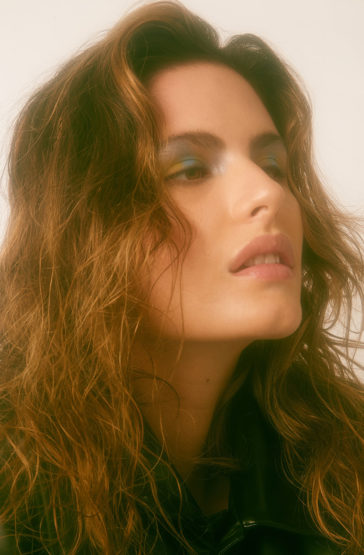Portrait of a woman with brown wavy hair and colorful eyeshadow, looking up, set against a soft, neutral background in a studio environment.