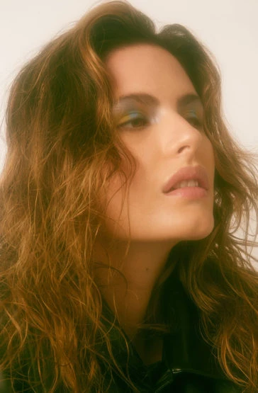 Portrait of a woman with brown wavy hair and colorful eyeshadow, looking up, set against a soft, neutral background in a studio environment.