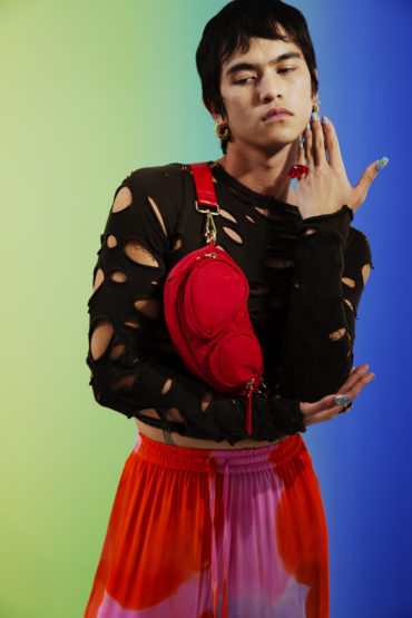 Person with red bag posing against a rainbow background. Wearing a black shirt with holes and orange and pink pants.