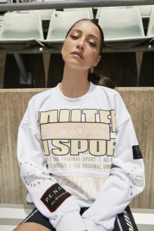 Woman in a white sweatshirt poses near stadium seating. The sweatshirt features the brand P.E Nation and has gold star details.